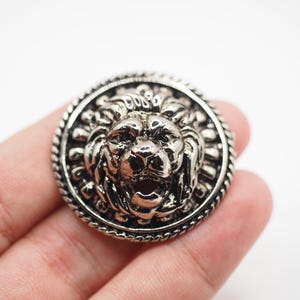 May include: Silver-toned lion head brooch with intricate detailing. The lion's face is centered within a circular frame, surrounded by a rope-like border. The brooch has a vintage or antique aesthetic.