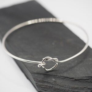 May include: A silver bangle bracelet with a knotted heart design. The bracelet is open and has a smooth, polished finish.