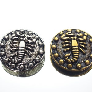 May include: Two round metal buttons, one silver, one bronze. Each has a detailed scorpion design in the centre, encircled by small, raised dots. These decorative buttons are likely for clothing or craft projects.