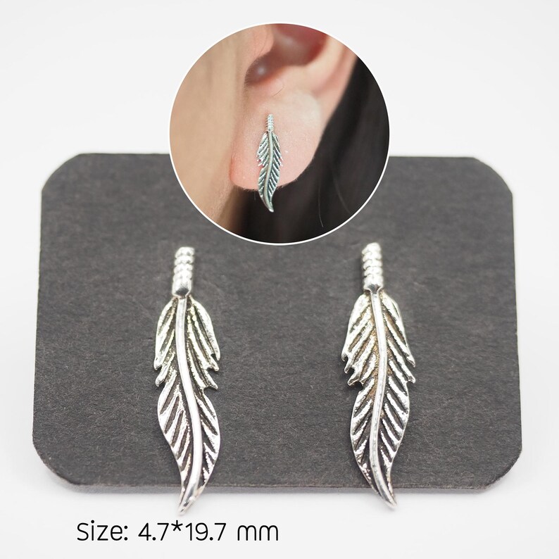 Puede incluir: Pendientes de pluma plateados. Los pendientes se muestran en una tarjeta negra. Los pendientes miden aproximadamente 1,97 cm de ancho y 4,7 mm de largo. Se muestra un pendiente puesto. Los pendientes tienen un dise&ntilde;o detallado de pluma.