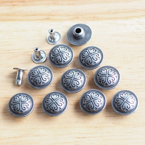 Op de afbeelding: Een verzameling antieke zilverkleurige metalen klinknagels en decoratieve knopen met een kruisachtig ontwerp. De knopen zijn rond en hebben een verhoogd patroon. De klinknagels hebben een pen- en kapontwerp. Op een licht houten oppervlak.