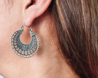 May include: Silver hoop earrings with an ornate, openwork design. The earrings have a crescent shape with detailed patterns and small beaded accents. The earrings are shown worn on an ear.