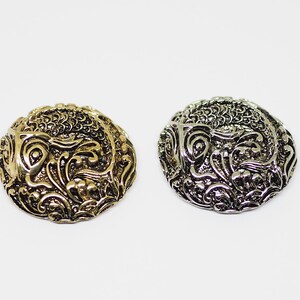 以下が含まれることがあります： 2つの装飾的な丸い金属製のボタン。1つは金色、もう1つは銀色です。どちらも、詳細な渦巻き模様の花柄とスクロールデザインが特徴です。ボタンは白い背景に配置されています。
