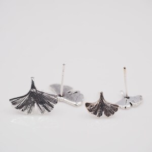 May include: Close-up of two pairs of silver-toned stud earrings. Each earring features a detailed ginkgo leaf design. The earrings are displayed on a white surface, showcasing their intricate details and metallic finish.