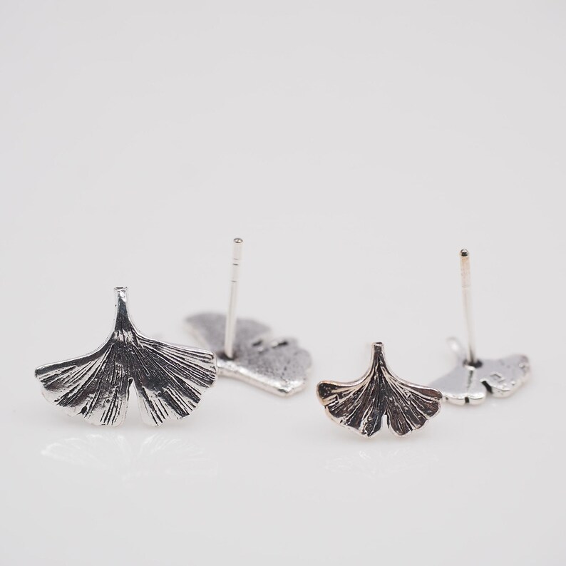 May include: Close-up of two pairs of silver-toned stud earrings. Each earring features a detailed ginkgo leaf design. The earrings are displayed on a white surface, showcasing their intricate details and metallic finish.