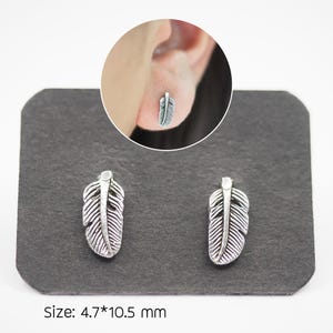 Puede incluir: Pendientes de pluma plateados. Cada pendiente presenta un dise&ntilde;o detallado de pluma. Los pendientes se muestran sobre una superficie gris oscuro. El tama&ntilde;o de los pendientes es de 4,7 mm x 10,5 mm.