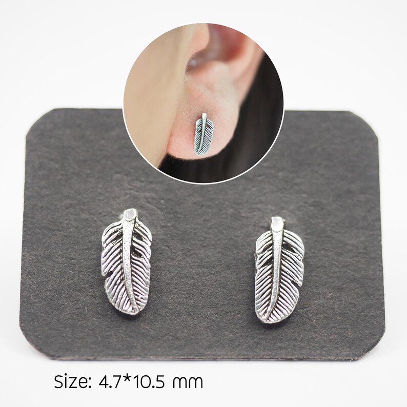 Puede incluir: Pendientes de pluma plateados. Cada pendiente presenta un dise&ntilde;o detallado de pluma. Los pendientes se muestran sobre una superficie gris oscuro. El tama&ntilde;o de los pendientes es de 4,7 mm x 10,5 mm.