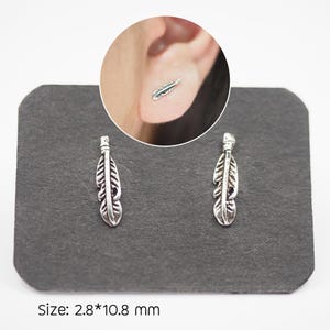 Puede incluir: Dos pendientes de bot&oacute;n plateados con forma de pluma, expuestos sobre una superficie gris oscuro. Los pendientes tienen l&iacute;neas y texturas detalladas, y un primer plano muestra uno en la oreja. El texto "Size: 2.8*10.8 mm" est&aacute; en la parte inferior. Medidas: 0,28 cm x 1,08 cm.