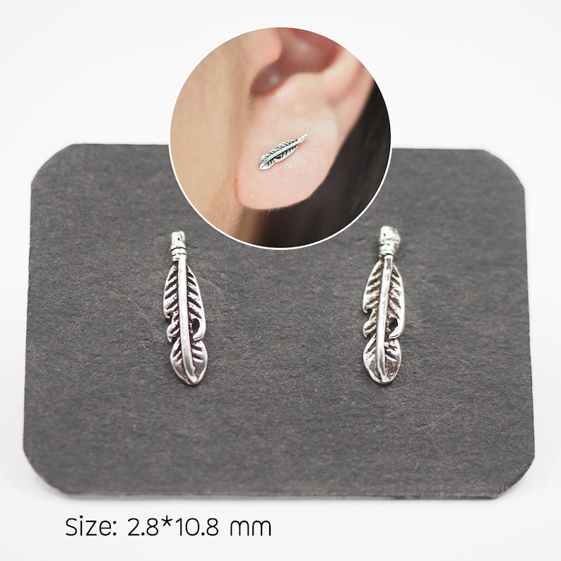 Puede incluir: Dos pendientes de bot&oacute;n plateados con forma de pluma, expuestos sobre una superficie gris oscuro. Los pendientes tienen l&iacute;neas y texturas detalladas, y un primer plano muestra uno en la oreja. El texto "Size: 2.8*10.8 mm" est&aacute; en la parte inferior. Medidas: 0,28 cm x 1,08 cm.