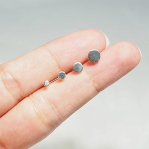 May include: Four silver stud earrings of different sizes, displayed on a finger.