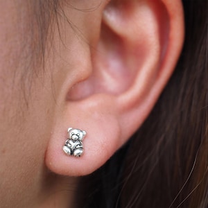 May include: A close-up of a silver teddy bear stud earring worn in a person's earlobe.