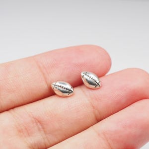 Könnte beinhalten: Zwei kleine, silberfarbene Football-förmige Charms. Jeder Charm hat detaillierte Stepplinien und eine strukturierte Oberfläche. Die Charms sind wahrscheinlich für die Schmuckherstellung oder das Basteln gedacht.