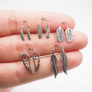 Puede incluir: Una colecci&oacute;n de pendientes plateados con forma de pluma, en varios dise&ntilde;os. Los pendientes se muestran en pares, con cinco estilos diferentes etiquetados del #1 al #5. Las plumas tienen texturas detalladas y est&aacute;n sobre un fondo claro.