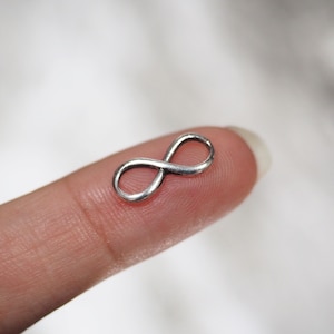 Può includere: Un piccolo ciondolo a forma di infinito color argento. Il ciondolo è tenuto su un dito, mostrando il suo design delicato. Il simbolo dell'infinito è un anello continuo, che rappresenta l'eternità e le infinite possibilità.
