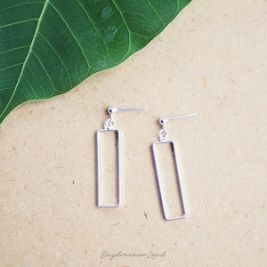 Könnte beinhalten: Ein Paar silberne, rechteckige Ohrringe mit Steckerverschluss. Die Ohrringe werden auf einer beigen Oberfläche mit einem großen grünen Blatt im Hintergrund präsentiert. Der Text "Daydreamer Land" ist unten zu sehen.