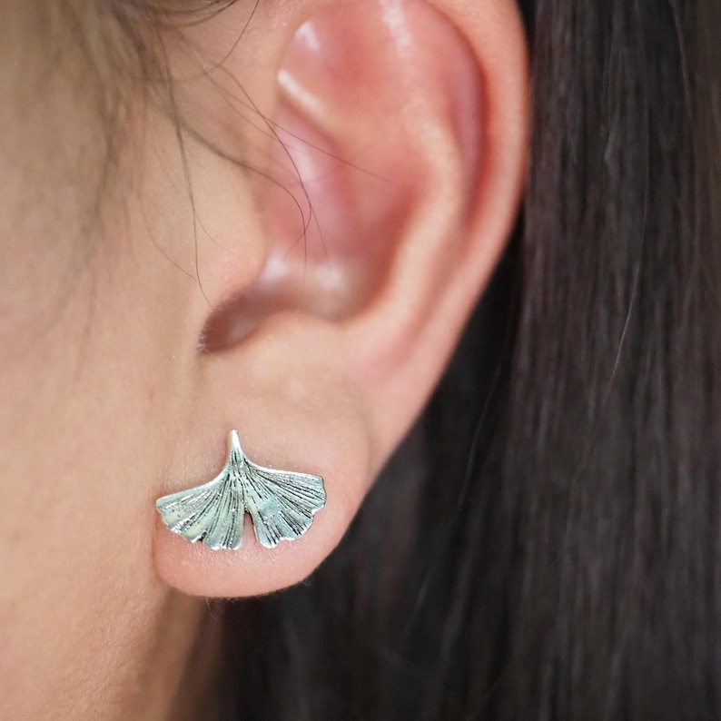 May include: Silver-colored ginkgo leaf stud earring. The earring is a detailed miniature replica of a ginkgo leaf, showcasing the leaf's distinctive fan shape and veining. The earring is worn on an ear.