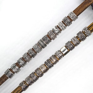 May include: Two wooden rods are adorned with numerous silver rings. The rings feature various designs, including swirls, geometric patterns, and floral motifs. The rods are brown, and the rings are silver, creating a contrast against a white background. The rings appear to be for hair.