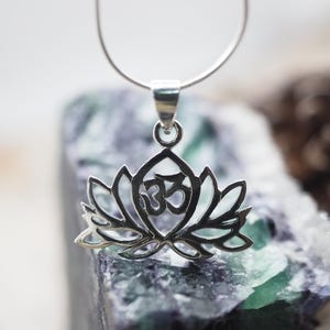 May include: A silver pendant necklace featuring a lotus flower design with an Om symbol in the center. The pendant is on a silver chain and is resting on a green and purple crystal.