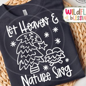 May include: A dark navy blue t-shirt with a white graphic design featuring a Christmas tree, snowflakes, and the text "Let Heaven & Nature Sing". The shirt is displayed on a woven, circular placemat.