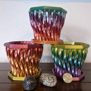 May include: Three colorful hexagonal planters with a woven design. The planters are stacked on top of each other, with the largest planter on the bottom and the smallest planter on top. The planters are painted in a gradient of colors, starting with green at the top and transitioning to purple at the bottom.