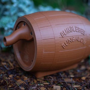 May include: A brown, barrel-shaped bumblebee house with the words "Bumblebee Bungalow" embossed on the side. The house has a spout-like entrance and is designed to provide shelter for bumblebees. It is made of a solid material and sits on a base.