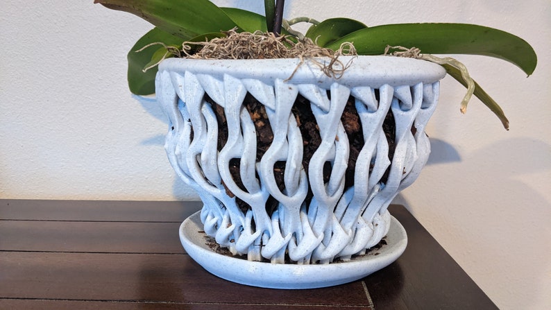 May include: A white ceramic orchid pot with a unique, woven design. The pot has a wide base and a narrow opening, and the woven design creates a lattice-like pattern. The pot is filled with soil and has a small saucer underneath.