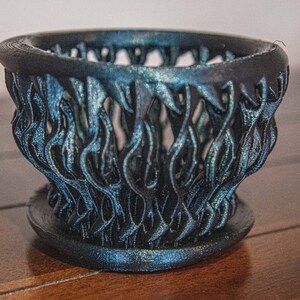 May include: A black and blue 3D printed planter with a unique, intricate design resembling flames. The planter has a wide base and a narrow opening.
