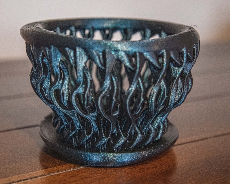 May include: A black and blue 3D printed planter with a unique, intricate design resembling flames. The planter has a wide base and a narrow opening.