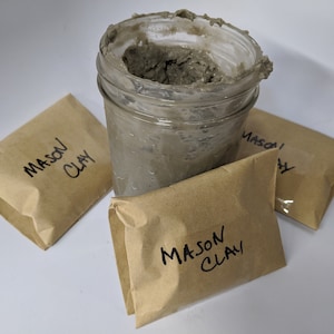 May include: Three brown paper bags labeled "MASON CLAY" and a glass jar filled with gray clay.