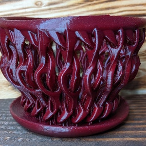 May include: A burgundy red 3D printed planter with a unique, intricate design resembling a woven basket. The planter has a wide, round base and a flared top.
