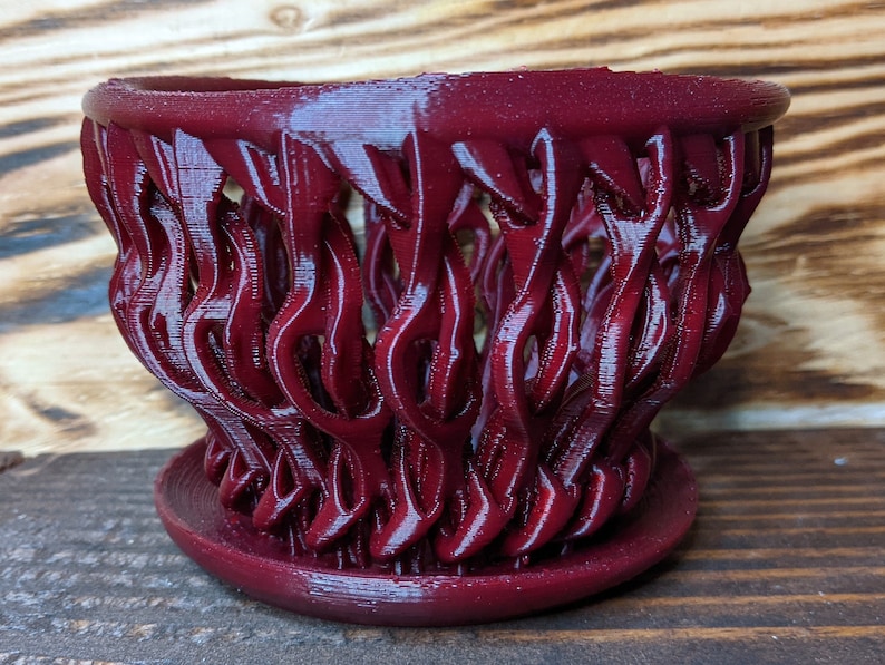 May include: A burgundy red 3D printed planter with a unique, intricate design resembling a woven basket. The planter has a wide, round base and a flared top.