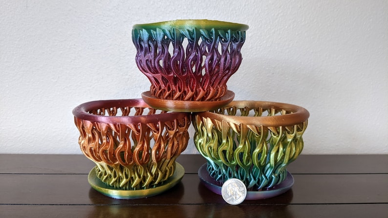 May include: Three rainbow-colored 3D printed plant pots, stacked on top of each other. The pots are made of a plastic-like material and have a unique, intricate design.