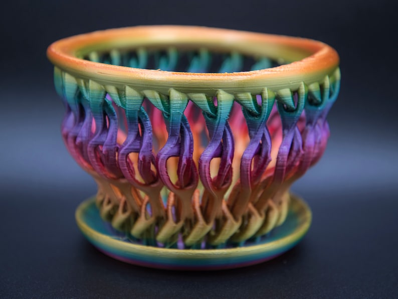 May include: A rainbow-colored, 3D printed planter with a decorative, open-weave design. The planter has a wide, round base and a narrow, cylindrical top.
