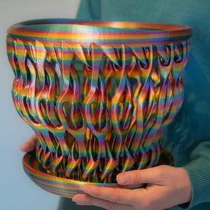 Könnte beinhalten: Ein regenbogenfarbener 3D-gedruckter Pflanztopf mit einem dekorativen, filigranen Design. Der Topf hat eine breite, offene Oberseite und eine kleinere Basis.