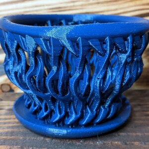 May include: A blue ceramic planter with a decorative, flame-like design. The planter has a wide, open top and a smaller base.