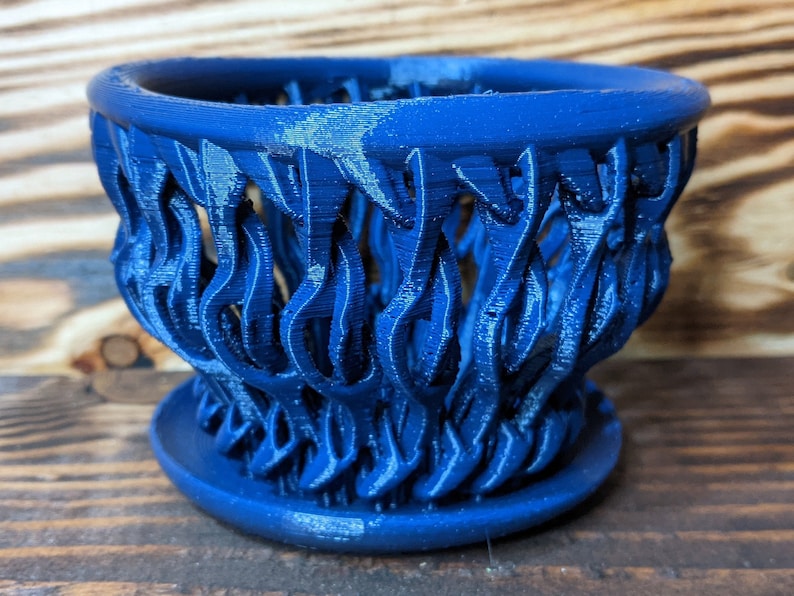 May include: A blue ceramic planter with a decorative, flame-like design. The planter has a wide, open top and a smaller base.
