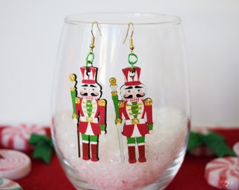 May include: A pair of dangle earrings featuring a red, white and green nutcracker design. The nutcracker is holding a stick and has a gold trim.