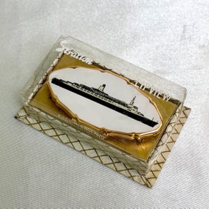 May include: A vintage Stratton compact mirror with a ship illustration in black and white. The compact is in a clear plastic case with gold-colored trim. The words "LIP VIEW" and "MADE IN ENGLAND" are visible on the packaging.