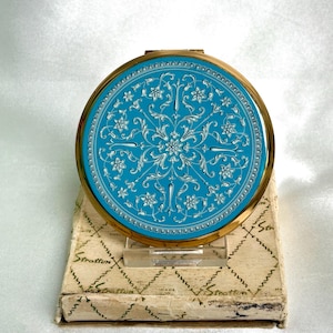 May include: A round gold-tone compact mirror featuring a bright turquoise enamel lid with an intricate white floral and scrollwork pattern. The compact rests on a rectangular off-white cardboard box with a repeating diamond pattern and the text Stratton.