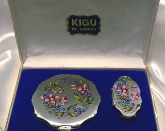 Op de afbeelding: Een Kigu of London vanity set in een crèmekleurige presentatiedoos met een blauwe fluwelen binnenkant. De set bevat een ronde compact en een kleinere ovale compact, beide met een zilverkleurige afwerking en een bloemmotief met roze bloemen.