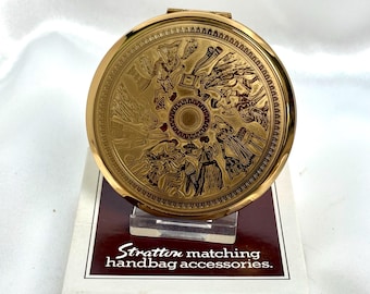 May include: A round gold-tone metal compact mirror featuring an intricate engraved scene of figures in classical attire. The compact rests on a white box with the text Stratton matching handbag accessories printed in dark brown.