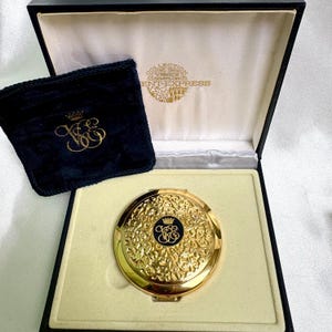 May include: A gold-toned compact mirror with an ornate floral design, presented in a black box with a cream-colored interior. A small, navy blue pouch with gold embroidery is also included. The compact features a black emblem with gold detailing.