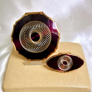 May include: A round compact mirror and a smaller oval compact mirror, both featuring a deep purple enamel finish with a gold-toned metal filigree overlay in a circular geometric pattern. Each piece has a scalloped gold-toned metal edge.
