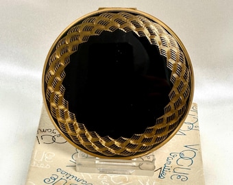 May include: A round gold-tone compact mirror featuring a black center inlay surrounded by a textured, woven-style gold border. The compact rests on a white box printed with the repeating blue text Vogue Vanities.