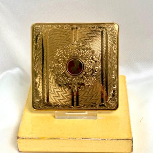 May include: A square gold-tone metal compact case featuring an intricate engraved pattern of radiating lines and floral borders. The center displays a circular medallion with a dark red inlay surrounded by a detailed embossed floral wreath.