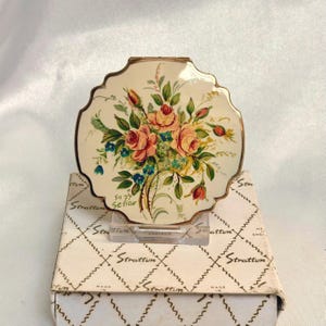 May include: A round, scalloped-edge Stratton powder compact with a cream-colored enamel finish featuring a painted floral bouquet of pink roses and small blue flowers. The compact is signed "Suzy Setiar" and rests on its original white box with a diamond pattern.