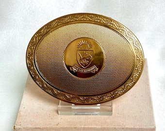 May include: An oval gold-toned compact mirror featuring a textured cross-hatch pattern on the surface and an ornate border. The center displays a circular emblem with a crest, a rising sun, and the engraved text "QUIS NOS SEPARABIT".