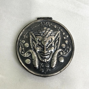 May include: A round, silver-toned compact with a hinged lid. The compact features a detailed, embossed design of a horned face with a smiling expression. The face is surrounded by decorative elements.