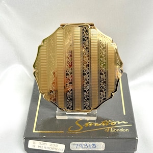 May include: A gold-toned, scalloped-edge compact mirror featuring vertical stripes with alternating textured and floral vine patterns. The compact rests on a dark grey box labelled Stratton of London with the text 79318 and 5 335 232 S MIRROR9/0.