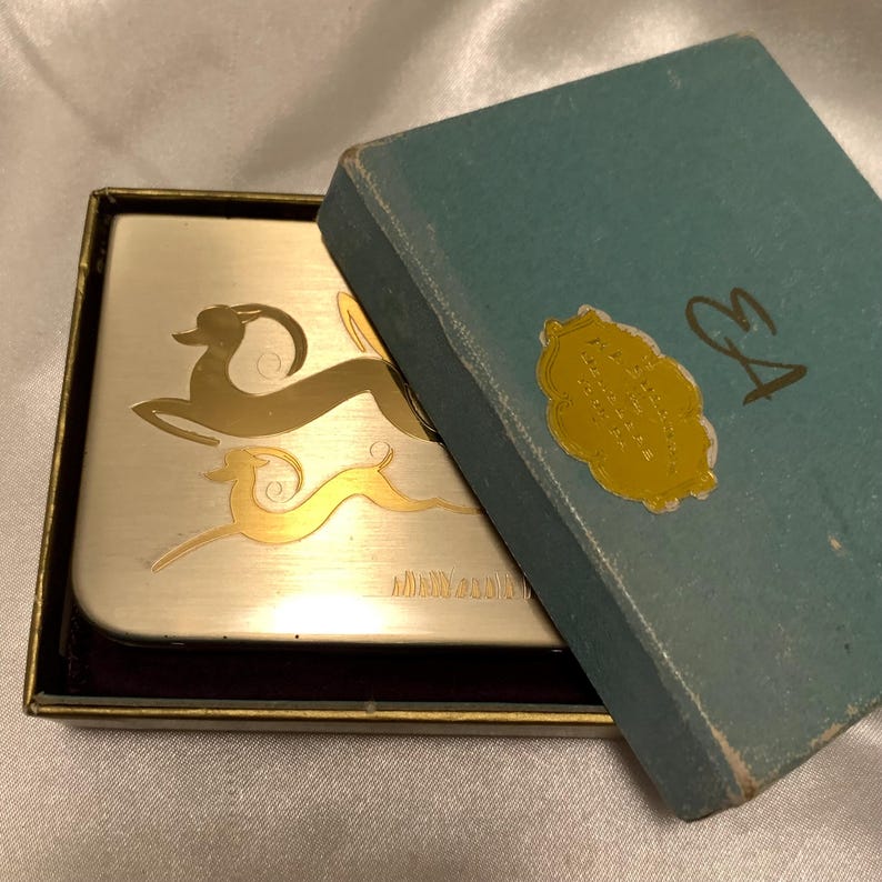 May include: A silver and gold compact with a design of two stylized deer. The compact is in a blue box with a gold label that reads "Elizabeth Arden New York".